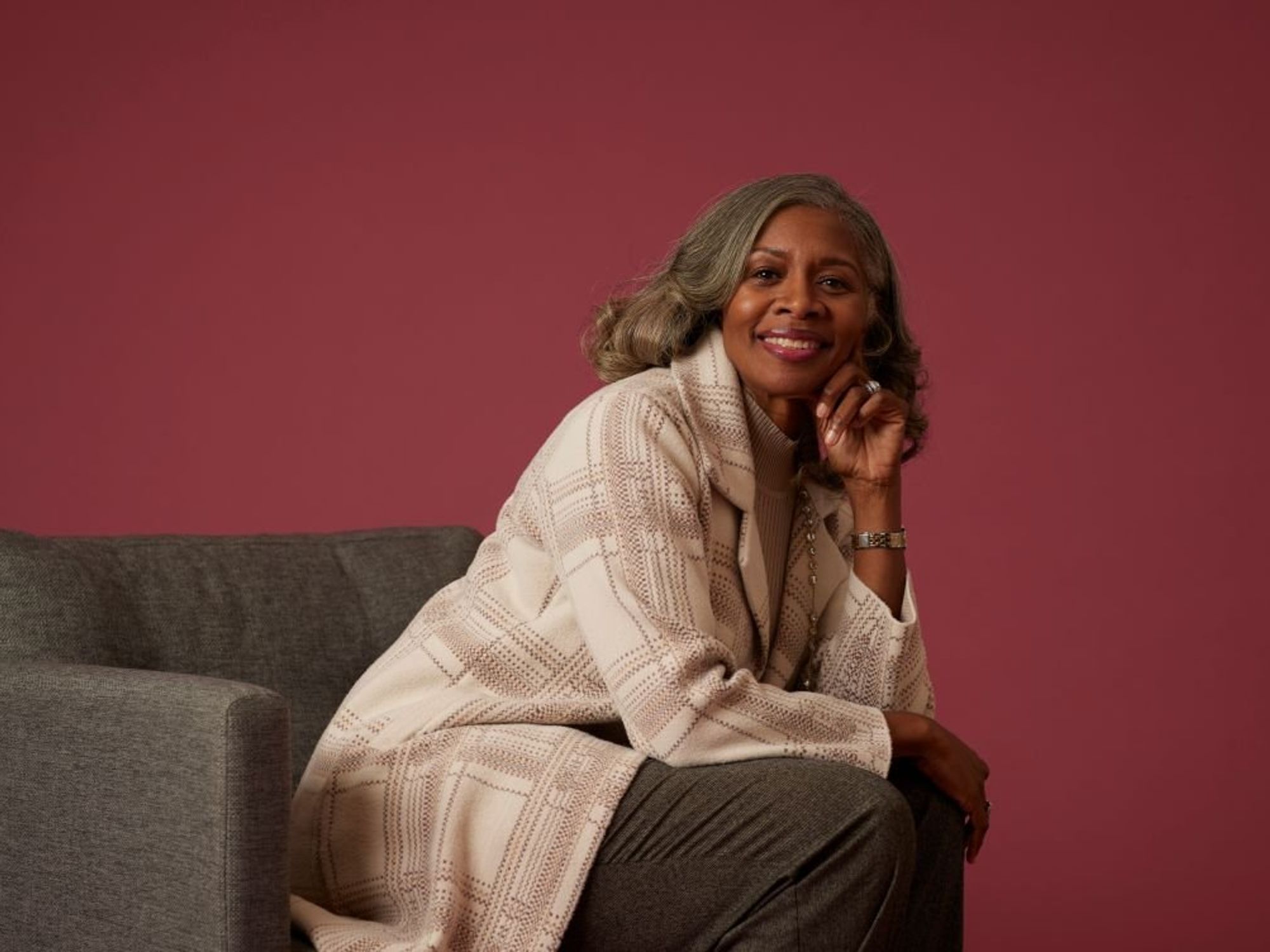 Smiling woman sitting on gray sofa, pink background.