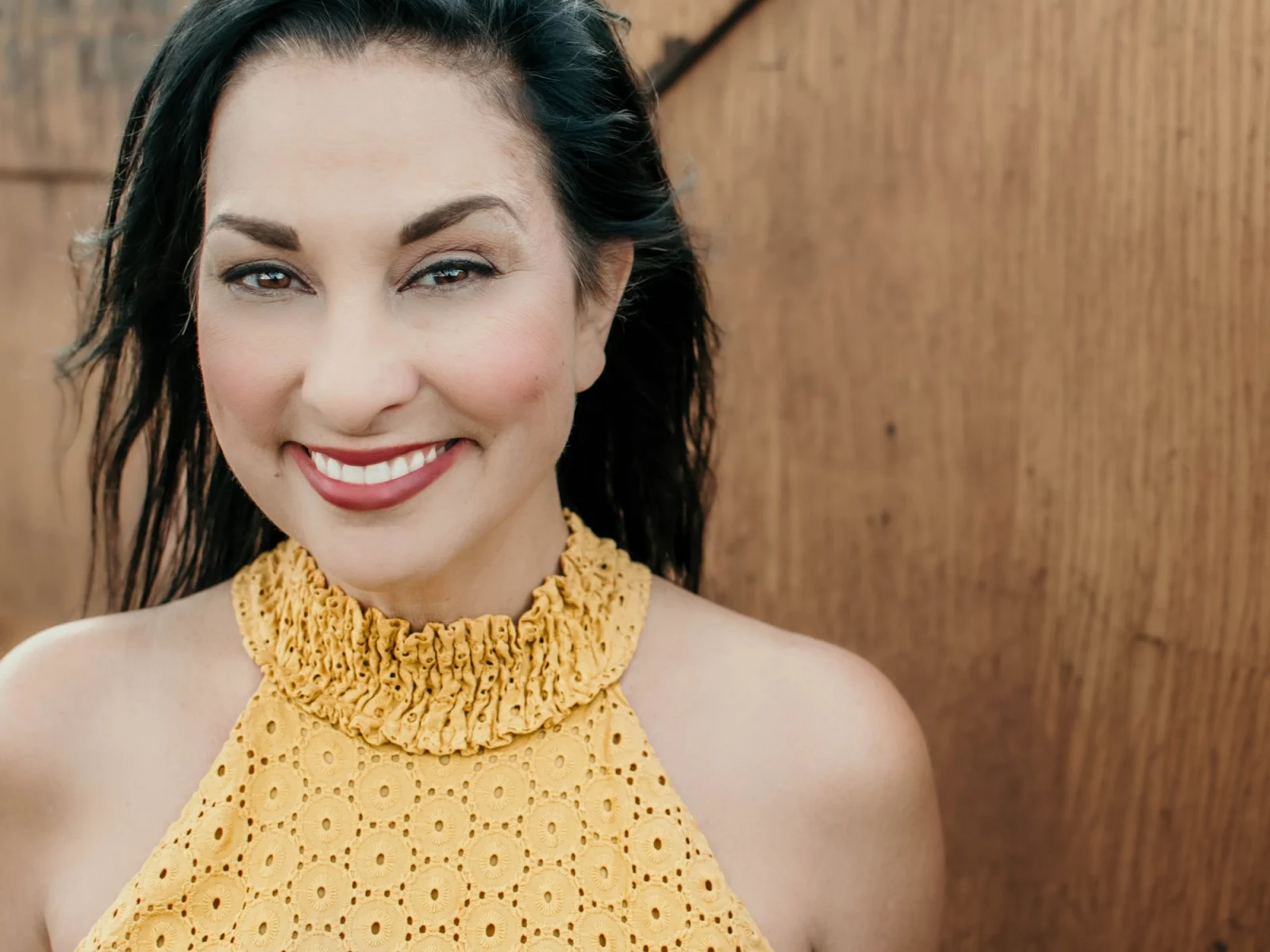 Smiling woman in yellow top, wooden backdrop.